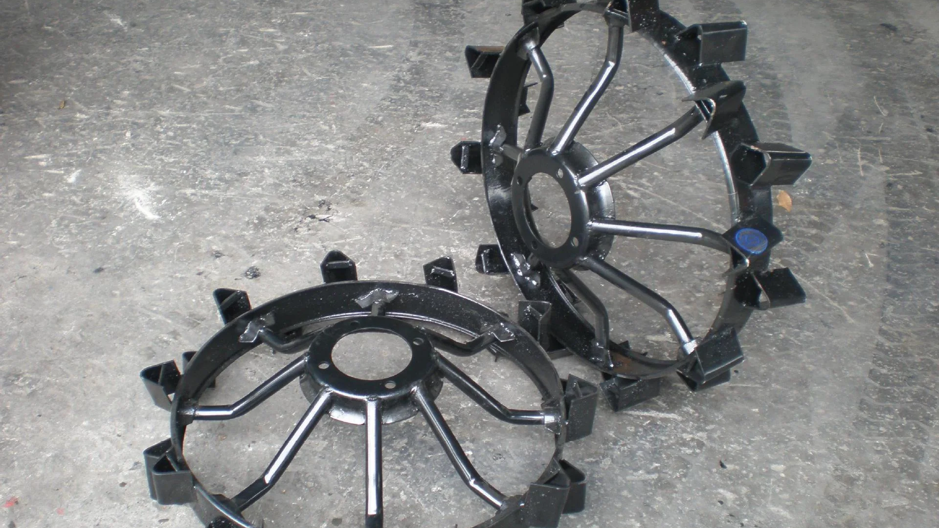 Two complete steel spiked wheels for a two-wheel tractor, lying on a workshop floor with their hub flanges, welded spokes and rim teeth in view