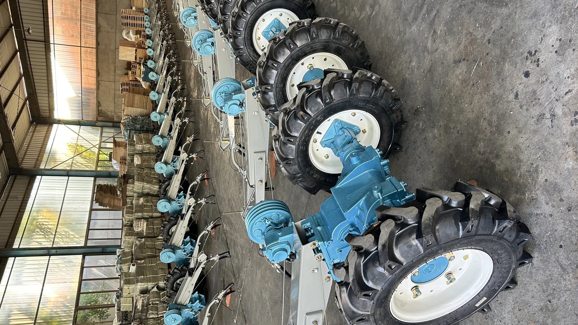 Rows of blue two-wheel tractors with their wheel assemblies fitted on a workshop floor, wooden export crates and wrapped implement sets alongside them