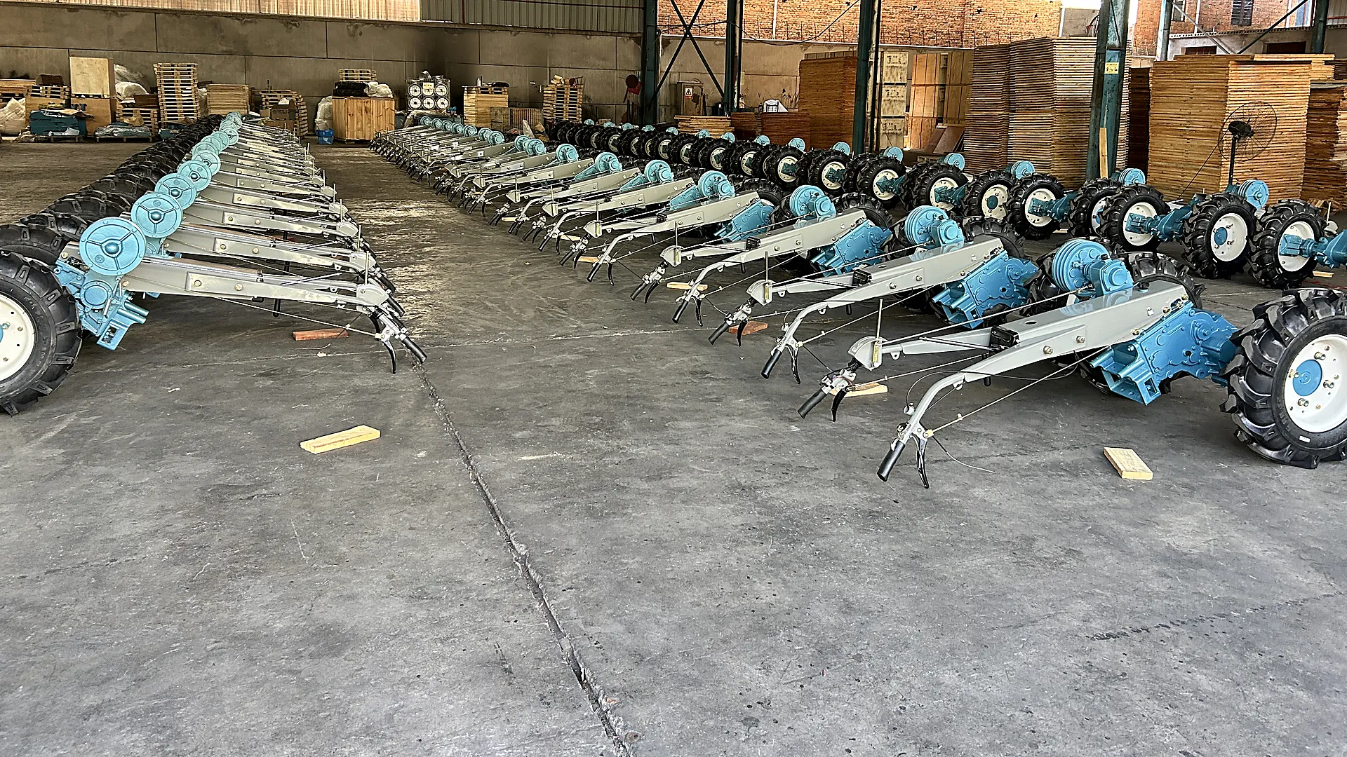Two long rows of complete two-wheel walking tractors on a workshop floor, with wooden export crates stacked behind them