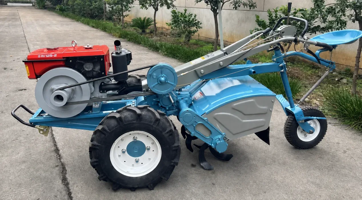 A blue two-wheel power tiller with its rotary tiller fitted, parked on a concrete apron with the curved tines hanging clear of the slab and the RV125-2 engine-model decal on the red cowl